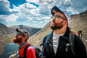 2 men outdoors on a mountainside, looking at sky wearing backpacks and NOZ sunscreen on nose