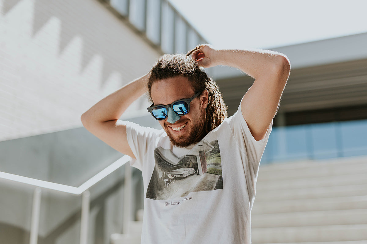 Guy smiling, wearing sunglasses, and a white shirt, pulling his hair back with blue NÖZ sun screen on nose